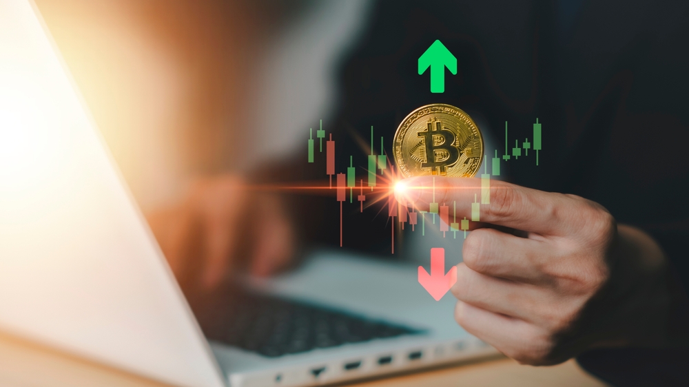 Bitcoin novčić ispred grafikona rasta i pada vrednosti na ekranu laptopa.